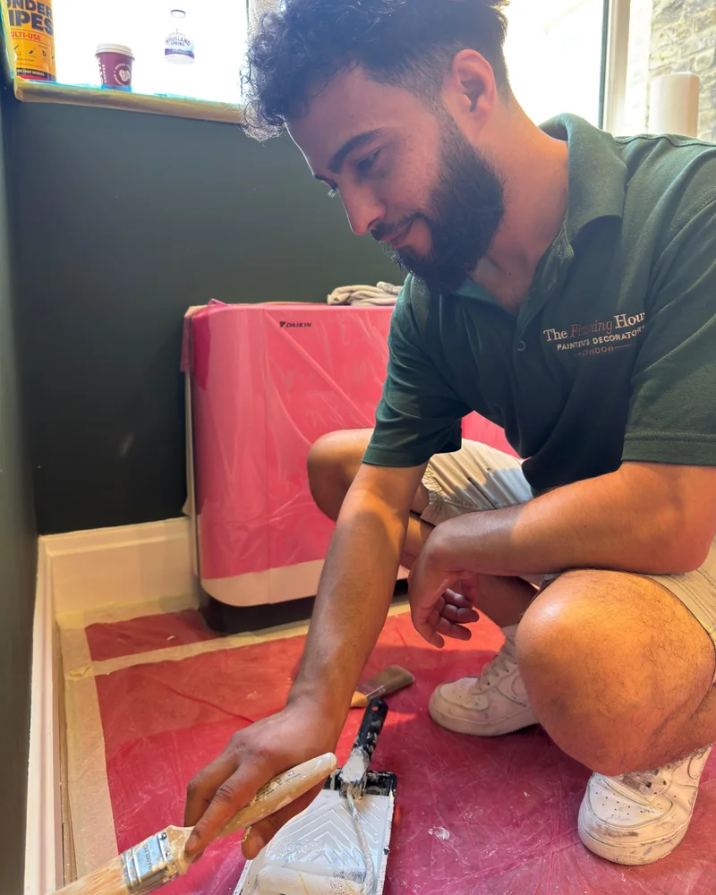 Decorator painting a skirting board by hand, with the floor and air-conditioning unit sheeted